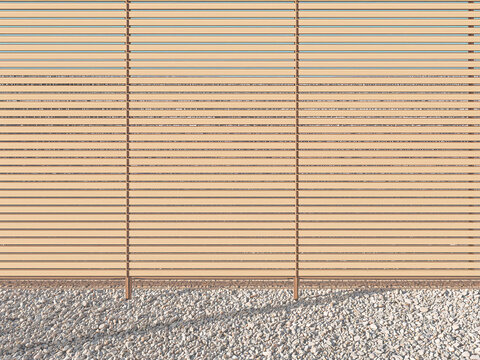 A horizontal wooden slatted fence panel with thin gaps positioned above a bed of light gravel with long shadows stretching across the ground surface