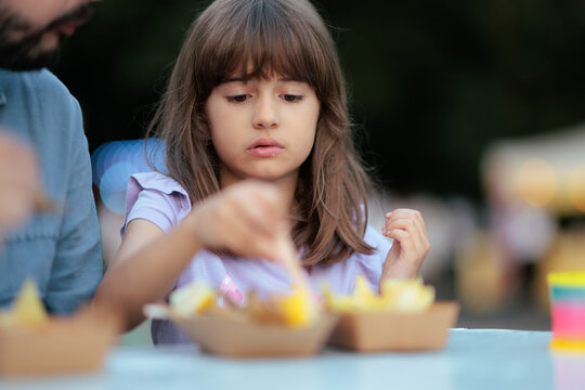 Cute Little Girl Eating French Fries From Paper Bowl Near Her Father. Focused young daughter enjoying delicious fast food chips during outdoor meal with parent