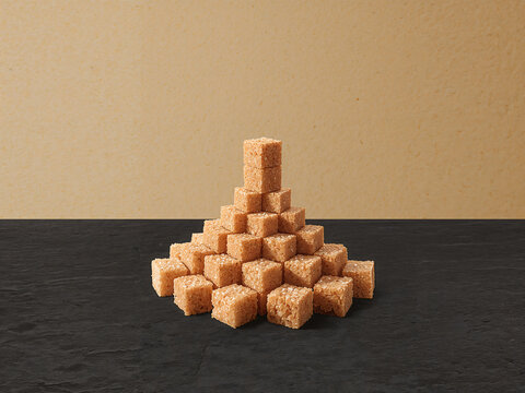 A pyramid stack of raw brown sugar cubes arranged on a dark textured surface against a solid light brown background, for food