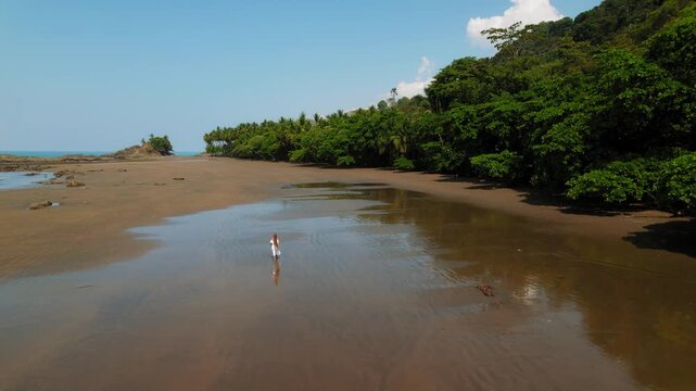 Woman in white dress walking on wide tropical beach near green forest on sunny day