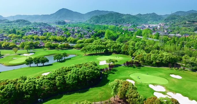 High angle view of a green golf course surrounded by lush trees, sand traps, small lakes, residential houses, and mountain peaks under bright sunlight.