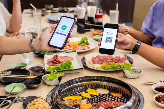 Selective focus to hands holding smartphones to scan a simulate QR code for contactless payment over a Japanese barbecue table at a restaurant.