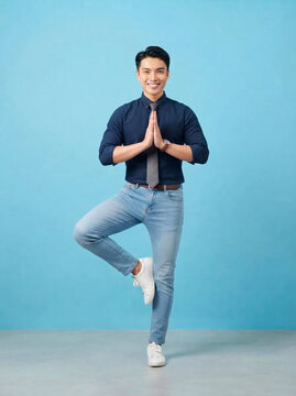 Businessman practicing yoga pose wearing formal shirt and tie while standing on one leg isolated against a solid light blue background in a professional studio