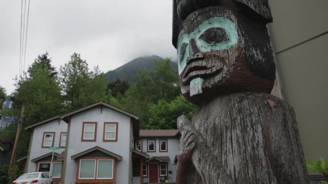 Weathered carved totem stands beside small houses on a cloudy day in an Alaska neighborhood street.