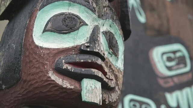 Weathered Alaska Native totem pole face fills the frame, showing carved details and faded turquoise paint against a soft-focus background designs.