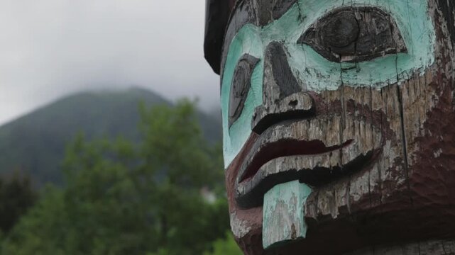 Close-up of a carved totem pole face in Alaska with a fog-covered green mountain in the background.