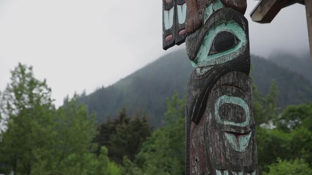 Weathered indigenous totem pole stands in front of misty tree-covered mountains on a cloudy day.