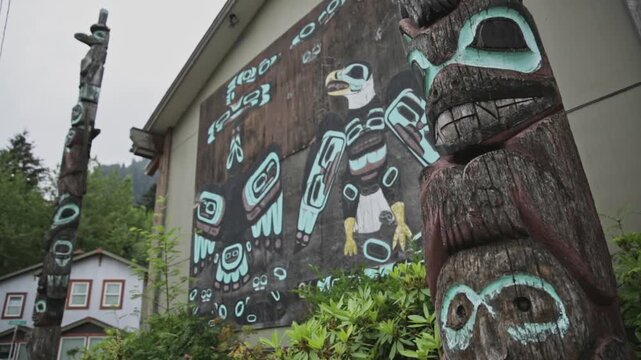 Carved totem poles frame a large Alaska Native clan painting on an exterior building wall in a forested neighborhood.