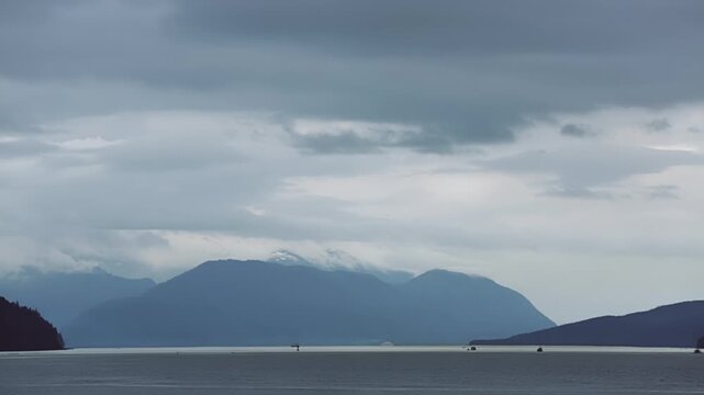 Layered blue hills fade into low clouds above a still Alaska fjord on an overcast day, with tiny boats barely visible on the water.