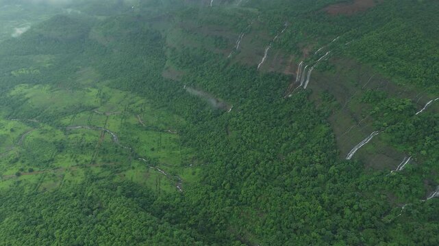 Aerial: Dolly reveal of multiple rainy season waterfalls plunging off sheer basalt wall into basin with low monsoon misty clouds