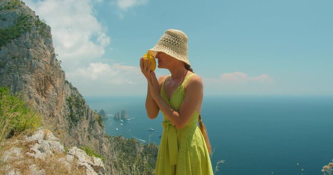 A traveler woman is inhaling the aroma of fragrant lemon used to make limoncello, on the background summer cliffs of Capri island with sea view