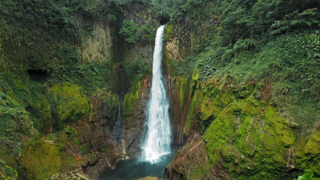 Surrounded by green cliffs and forest, a tall waterfall flows dramatically into a clear pool in Costa Rica