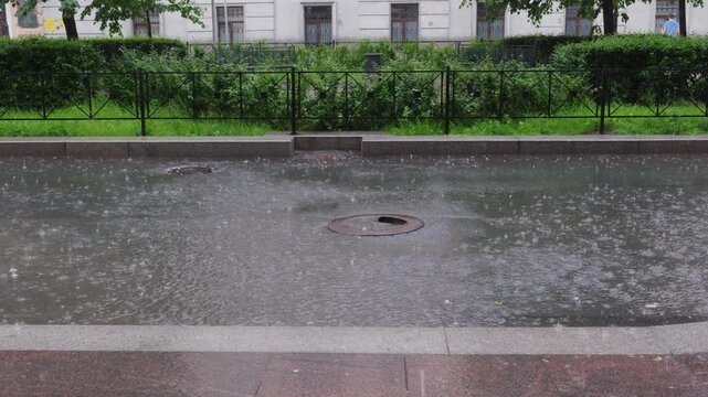 Stationary shot captures downpour striking shallow waterway with metal sewer grate, bordered by lush green hedge, classical architecture visible beyond, overcast natural lighting