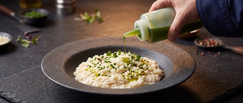 Professional chef's hand squeezes vibrant green herbal oil over creamy white risotto in elegant dark bowl