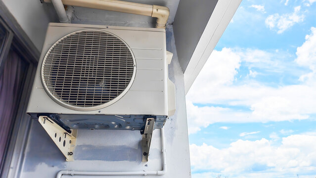 Air Conditioning Compressor Unit on a Building Exterior: Low angle view of split system air conditioner condenser unit outdoors, with a clear blue sky and fluffy clouds visible in the background.