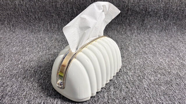 A modern aesthetic white plastic tissue box dispenser with a fluted, shell-like design, clear acrylic handle, gold accents, and one tissue pulled out, resting on a grey textured background