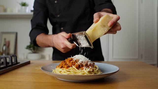 Cook grates hard cheese over pasta with meat sauce in a fixed kitchen shot