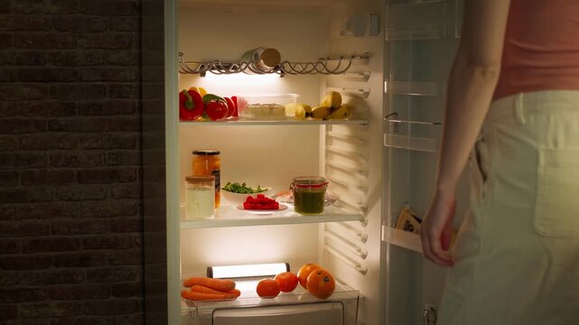 Woman standing in front of an open refrigerator, selecting and taking ingredients from inside before closing the fridge in a cozy home kitchen interior.