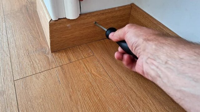 Securing a wooden baseboard to the wall with a screwdriver, completing laminate floor installation and interior finishing during home renovation and repair work