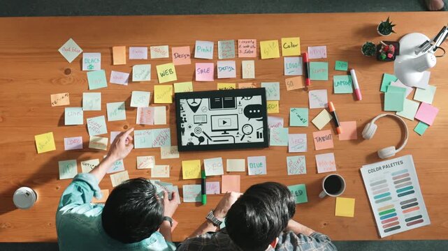 Top view of creative designer working together brainstorming idea by using sticky notes at meeting table with color palettes. Aerial view of smart team choosing icon graphic from tablet. Symposium.