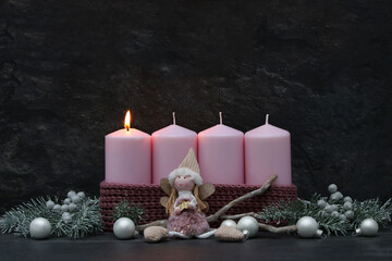 Fototapeta premium First Sunday of Advent: A burning pink candle on a modern Advent wreath featuring an angel figure, fir branches, and silver baubles against a dark background, with copy space.