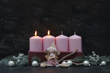 Fototapeta premium Second Sunday of Advent: Two burning pink candles on a modern Advent wreath featuring an angel figure, fir branches, and silver baubles against a dark background, with space for text.