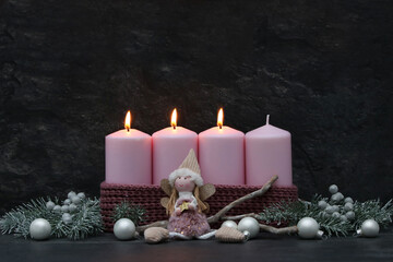 Fototapeta premium Third Sunday of Advent: Three burning pink candles on a modern Advent wreath featuring an angel figure, fir branches, and silver baubles against a dark background, with copy space.