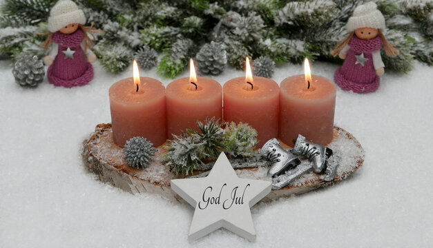 Four burning candles on a wooden board with pine cones, ice skates, and a star bearing the inscription "God Jul" in the snow, surrounded by two small figures wearing hats and scarves.
