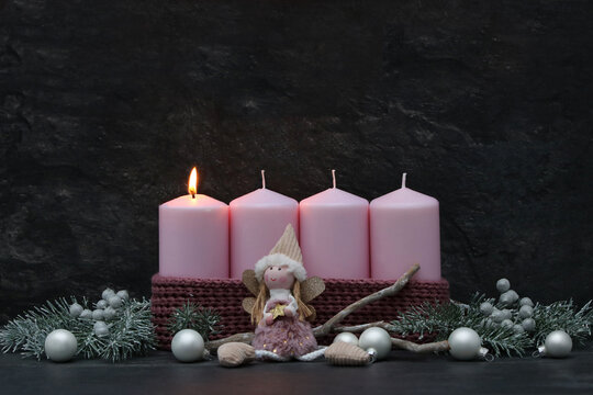 First Sunday of Advent: A burning pink candle on a modern Advent wreath featuring an angel figure, fir branches, and silver baubles against a dark background, with copy space.