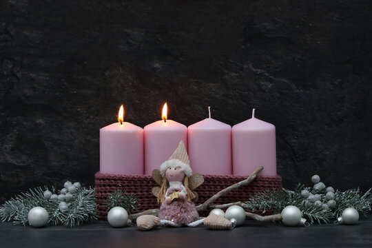 Second Sunday of Advent: Two burning pink candles on a modern Advent wreath featuring an angel figure, fir branches, and silver baubles against a dark background, with space for text.