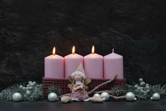 Third Sunday of Advent: Three burning pink candles on a modern Advent wreath featuring an angel figure, fir branches, and silver baubles against a dark background, with copy space.