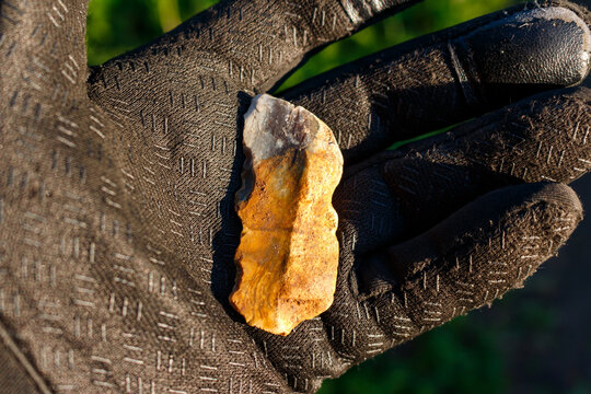 Rough textured flint stone shaped like an ancient prehistoric scraper held in a protective work glove, close-up archaeological find against a blurred natural outdoor background