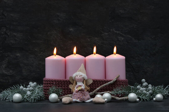 Fourth Sunday of Advent: Four burning pink candles on a modern Advent wreath with an angel figurine, fir branches, and silver baubles against a dark background, with space for text.