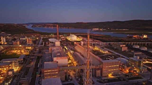 The view shows an industrial area with various buildings and equipment near a river during dusk. Lights are on, indicating ongoing activity. Vessels can be seen in the water.