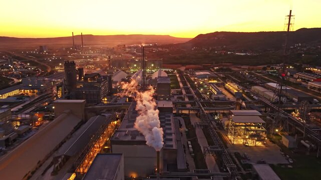 Smoke rises from a factory in an industrial area as the sun sets in the background. The lights of buildings begin to shine in the early evening.