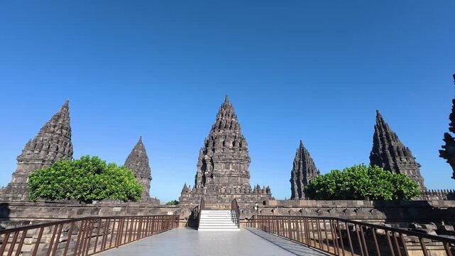 Symmetrical pathway leads toward the towering Prambanan Temple complex through landscaped gardens under a cloudless blue sky.