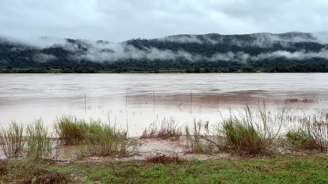 Timelapse video showing the wide brown river flowing past green mountains covered in low thick fog. Beautiful scenic landscape of tropical wilderness on a cloudy rainy day.