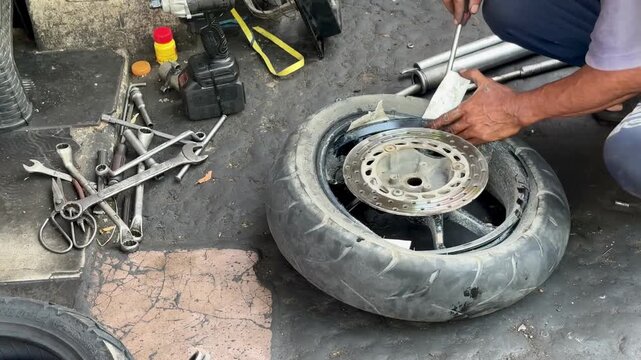 An Asian man is removing a tire from the rim using a manual pry bar, with a piece of thick paper to prevent damage to the rim