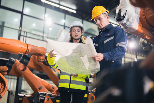 Engineers reviewing a technical blueprint beside industrial robotic arms in a factory, manufacturing automation, production planning, engineering collaboration, and equipment installation