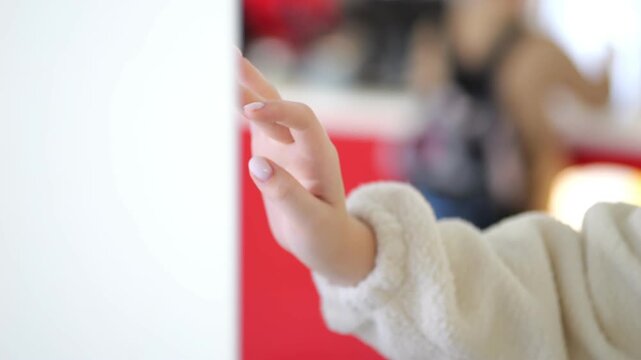 Close-up hand touches white surface. Person wears cream fleece sleeve. Background shows blurred red interior. Warm lighting creates soft focus effect
