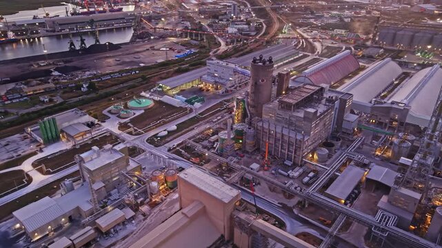 A bird's eye view shows a large industrial complex with various buildings, machinery, and roads. The scene captures the layout and activity around the site during the evening.