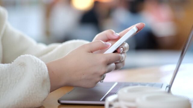 Person holds phone, laptop beside. Warm cafe setting, blurred background. Hands in soft sweater, coffee cup nearby. Focus on modern device usage, relaxed atmosphere