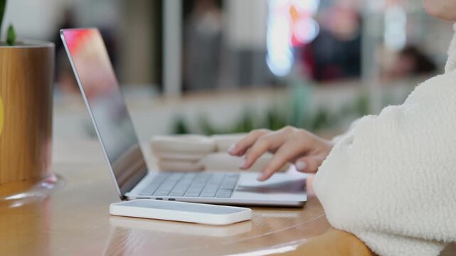 Person types on laptop at wooden table. Warm lighting, cozy sweater, blurred background. Focus on hands and keyboard. Coffee cup nearby. Office or cafe setting