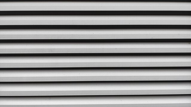 A modern industrial louvered vent in light gray, featuring a repeating geometric pattern of parallel horizontal lines and dark shadows.
