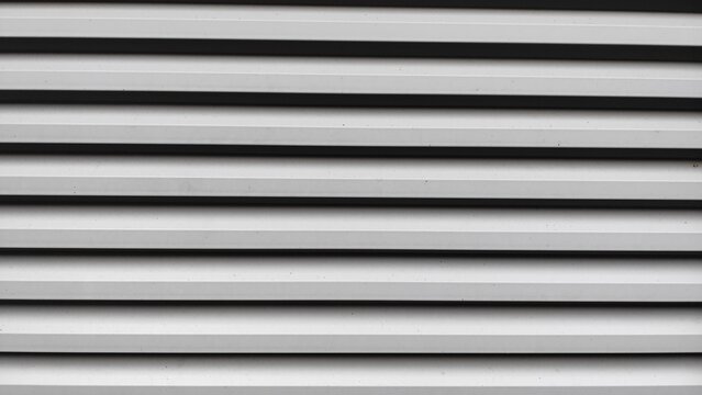 A modern industrial louvered vent in light gray, featuring a repeating geometric pattern of parallel horizontal lines and dark shadows.
