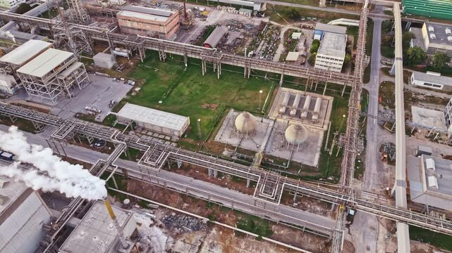Aerial view shows an industrial site with pipes, storage tanks, and vegetation. The layout includes multiple structures and roads with visible activity during the day.