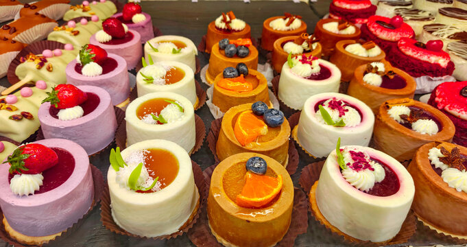 Assorted miniature gourmet cakes sit displayed in a professional bakery showcase. These colorful desserts feature fresh fruit toppings, delicate cream, and berry glazes