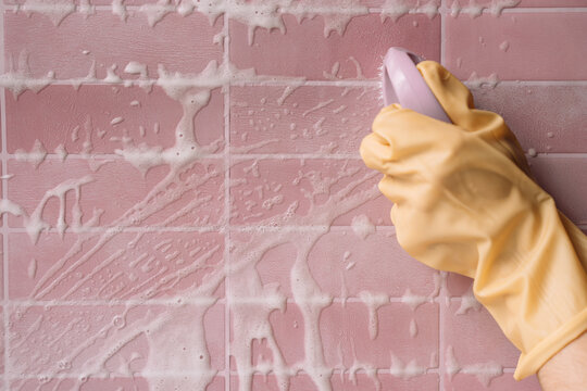 Gloved hand scrubbing pink tiles with a purple brush through white cleaning foam.