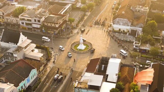 Aerial drone footage of Tugu Yogyakarta at a busy urban intersection, surrounded by traffic, roads, buildings, and warm golden morning sunlight in Yogyakarta, Indonesia.