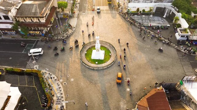 Aerial drone footage of Tugu Yogyakarta at a busy urban intersection, surrounded by motorcycles, cars, roads, and city buildings during warm morning light in Yogyakarta, Indonesia.